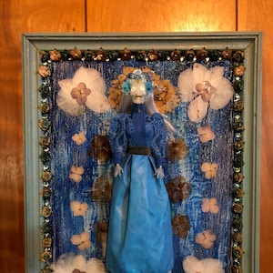 May include: A framed artwork featuring a doll with a blue dress and a floral crown, surrounded by dried flowers on a blue background.