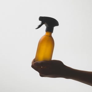 Könnte beinhalten: Eine Hand hält eine braune Glas-Sprühflasche mit einem schwarzen Plastiksprühkopf. Die Flasche ist matt und hat eine matte Oberfläche.