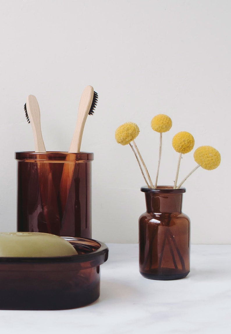 Puede incluir: Dos cepillos de dientes de madera con cerdas negras se colocan de pie en un frasco de vidrio marr&oacute;n. Un peque&ntilde;o frasco de vidrio marr&oacute;n con un ramo de flores secas amarillas se encuentra sobre una superficie blanca.