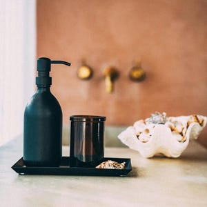 Puede incluir: Una botella de dispensador de jab&oacute;n negro con una bomba, un tarro de vela de vidrio marr&oacute;n y una concha de mar blanca en una bandeja negra.