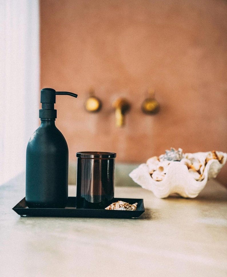 Puede incluir: Una botella de dispensador de jab&oacute;n negro con una bomba, un tarro de vela de vidrio marr&oacute;n y una concha de mar blanca en una bandeja negra.