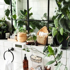 Puede incluir: Un lavabo de ba&ntilde;o blanco con un toallero de metal negro, una botella de jab&oacute;n de vidrio marr&oacute;n, una cesta de alambre negro con esponjas blancas, un tarro de vidrio marr&oacute;n con un cepillo de dientes blanco y un tarro de vidrio marr&oacute;n con un cepillo blanco. El lavabo est&aacute; rodeado de azulejos blancos y un espejo enmarcado en negro con plantas reflejadas en &eacute;l.