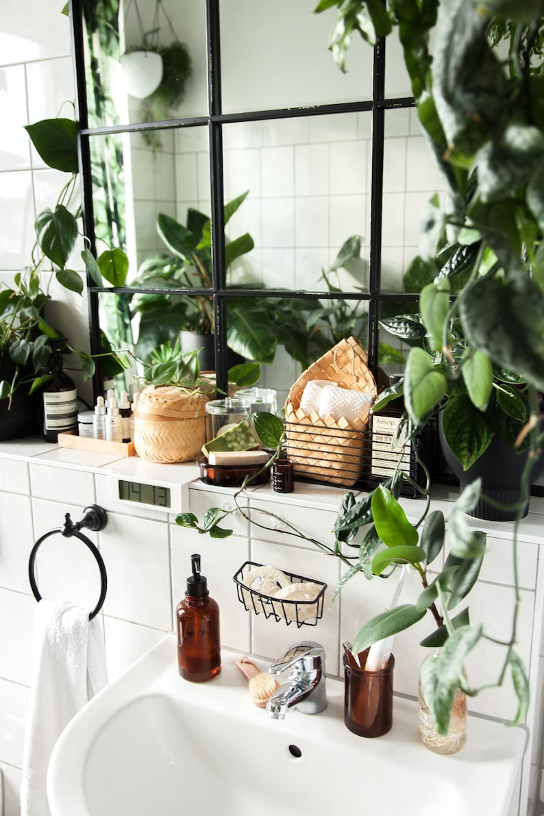 Puede incluir: Un lavabo de ba&ntilde;o blanco con un toallero de metal negro, una botella de jab&oacute;n de vidrio marr&oacute;n, una cesta de alambre negro con esponjas blancas, un tarro de vidrio marr&oacute;n con un cepillo de dientes blanco y un tarro de vidrio marr&oacute;n con un cepillo blanco. El lavabo est&aacute; rodeado de azulejos blancos y un espejo enmarcado en negro con plantas reflejadas en &eacute;l.