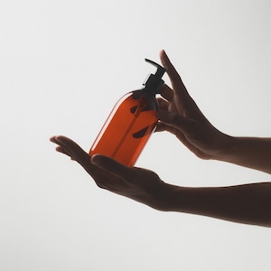 May include: A hand holds a brown amber glass bottle with a black pump dispenser. The bottle is filled with a clear liquid.