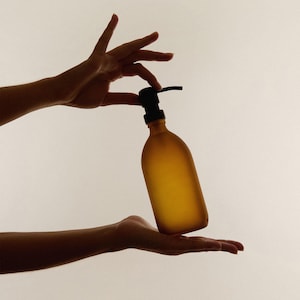May include: A frosted amber glass bottle with a black pump dispenser. The bottle has a rounded, cylindrical shape and a smooth, matte texture. The pump mechanism is positioned at the top, featuring a black nozzle and a circular base collar.