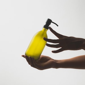 May include: A person's hands holding a yellow glass bottle with a black pump dispenser.