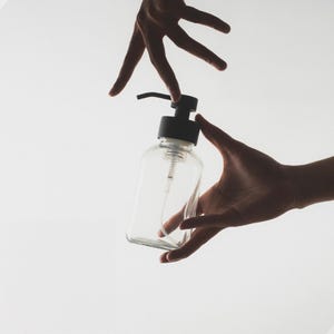 May include: A clear glass bottle with a black pump dispenser. The bottle is empty and has a textured surface.