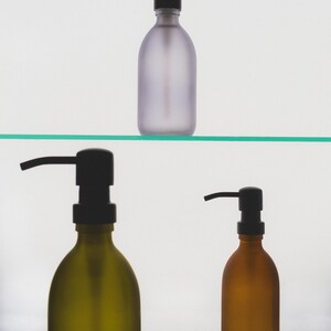 May include: Three frosted glass bottles with black pump tops. The bottles are on a glass shelf. The bottles are different colours: purple, green, and brown.