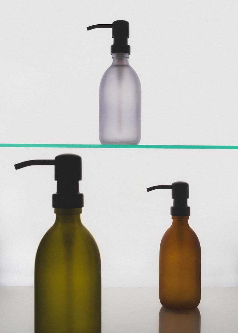 May include: Three frosted glass bottles with black pump tops. The bottles are on a glass shelf. The bottles are different colours: purple, green, and brown.