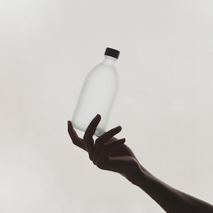 May include: A frosted glass bottle with a black lid is held by a hand against a white background.