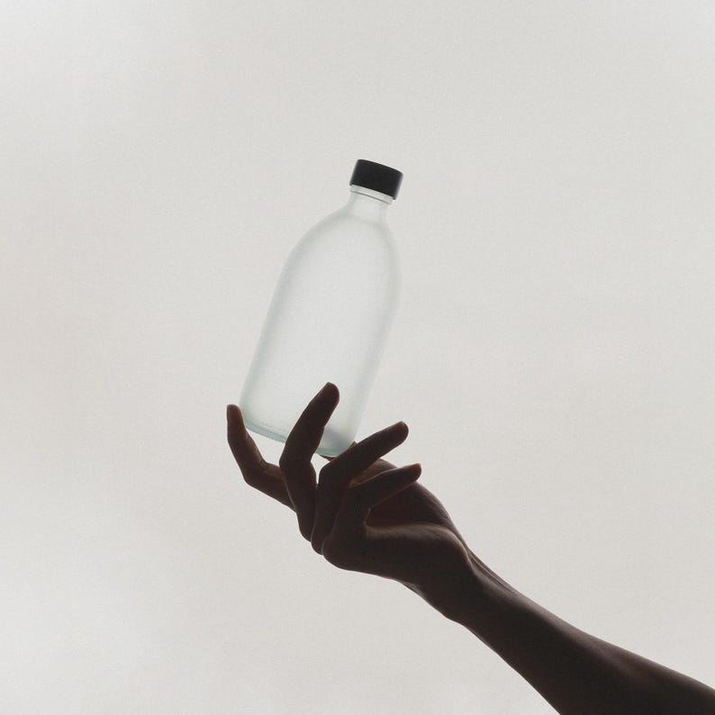 May include: A frosted glass bottle with a black lid is held by a hand against a white background.