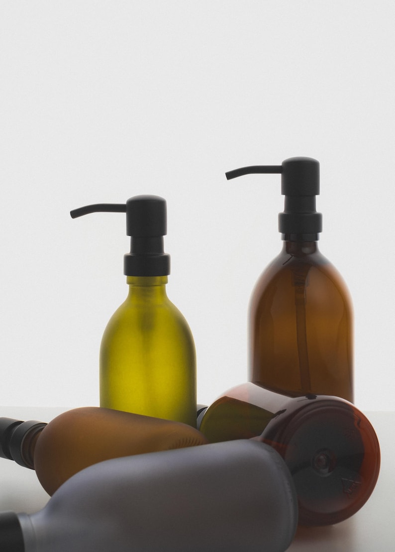May include: Four glass bottles with black pump tops. Two bottles are amber coloured, one is green, and one is brown. The bottles are on a white surface.