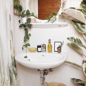 May include: A white bathroom sink with a chrome tap, a yellow soap dispenser, a black candle, and a small yellow bowl. The sink is surrounded by white tiles and green plants.
