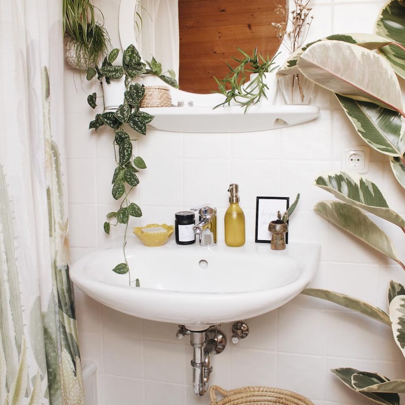 May include: A white bathroom sink with a chrome tap, a yellow soap dispenser, a black candle, and a small yellow bowl. The sink is surrounded by white tiles and green plants.