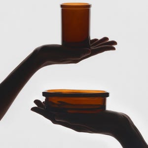 Puede incluir: Dos recipientes de vidrio &aacute;mbar, uno alto y cil&iacute;ndrico, el otro un cuenco poco profundo, se sostienen en manos extendidas.