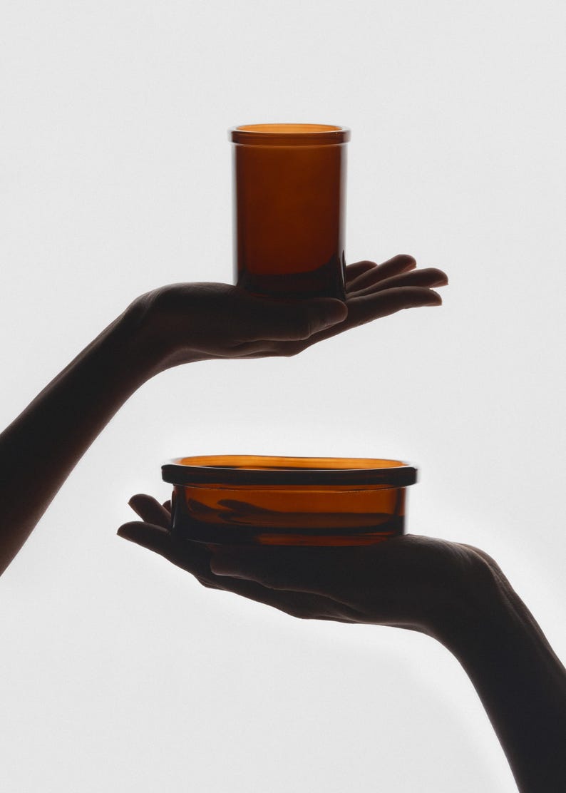 Puede incluir: Dos recipientes de vidrio &aacute;mbar, uno alto y cil&iacute;ndrico, el otro un cuenco poco profundo, se sostienen en manos extendidas.