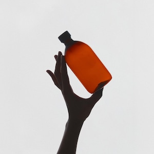 Può includere: Una bottiglia colorata di arancione con un tappo nero è tenuta su uno sfondo bianco. La bottiglia ha una forma rettangolare con bordi arrotondati. La sagoma di una mano è visibile, che tiene la bottiglia.