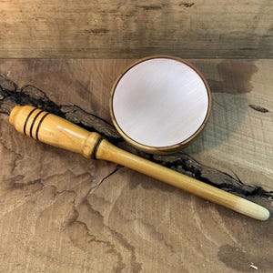 May include: A wooden mallet with three black stripes near the head and a round, light-colored, metallic-looking object with a wooden rim. The mallet is light brown and the round object is light pink.