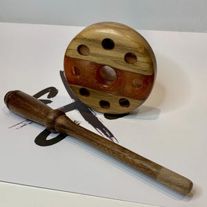 May include: A wooden pipe stand with a dark brown handle and a round base. The base has a reddish-brown center and several holes. The handle is long and slender, with a lighter-colored tip. The stand is made of wood.