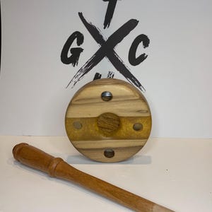 May include: A wooden pipe stand and matching pipe. The stand is circular, featuring light wood grain and a gold resin inlay. The pipe is a similar colour, with a light-coloured tip. The background displays the letters "GTC" in a black design.