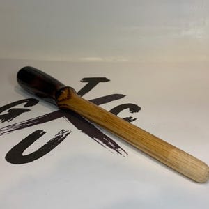 May include: A wooden tool with a dark brown handle and a light brown body. The tool has a rounded end and a slightly thicker section where the handle meets the body. The tool is lying on a white surface with black text.