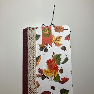 May include: A small, white notebook with a fall-themed design. The cover features a variety of leaves, acorns, and a floral bouquet. The notebook is decorated with lace, plaid fabric, and a paper banner that says "Gratitude".