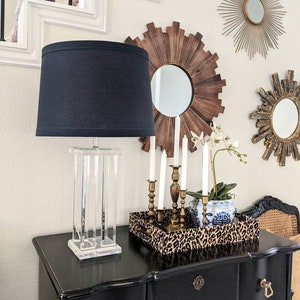 May include: A black lamp with a clear acrylic base sits on a black dresser with gold hardware. The lamp is next to a tray with a leopard print design, a white and blue porcelain vase with a floral arrangement, and a set of gold candle holders with white candles.