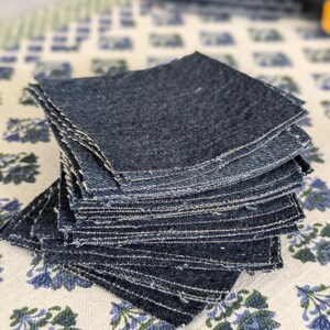 May include: A stack of dark blue denim fabric squares with frayed edges. The squares are stacked on top of each other, with a few more squares visible in the background. The fabric has a textured appearance.