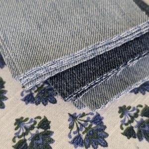 May include: Close-up of denim fabric samples in varying shades of blue. The samples are stacked, showcasing the texture and color variations. Below, a white fabric with a blue floral pattern adds contrast.