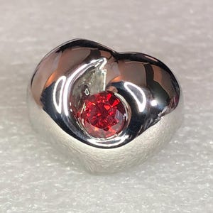 May include: A silver-toned ring with a heart-shaped design. The ring features a vibrant, round, red gemstone set in the center. The metal has a polished, reflective surface, creating a striking contrast with the red stone.