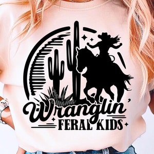 May include: A light peach-colored t-shirt featuring a black graphic of a cowgirl riding a horse, cacti, and the words "Wranglin' FERAL KIDS." The design is set within a semi-circle. The text "Crafty Mama Studios" is also visible.