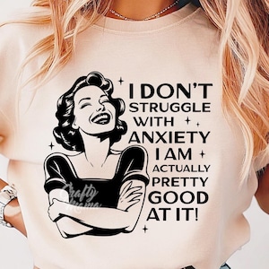 以下が含まれることがあります： 薄いピンクのTシャツに、腕を組んだ女性の白黒のグラフィックがプリントされています。グラフィックには、「私は不安と闘っていません。実際、私はとても得意なんです！」というテキストが含まれています。