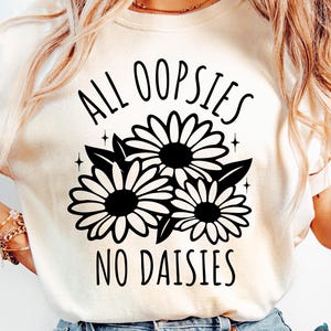 May include: White t-shirt with black text that reads "All oopsies no daisies" and a black graphic of three daisies.