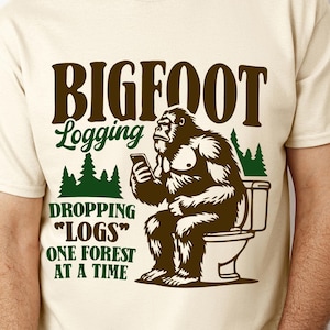 May include: Cream-colored t-shirt with a humorous graphic featuring a Bigfoot character on a toilet, looking at a phone. The text reads "BIGFOOT Logging" and "DROPPING 'LOGS' ONE FOREST AT A TIME" in brown and green.