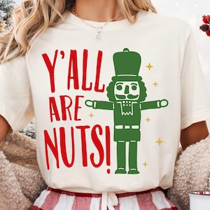 May include: Cream-coloured t-shirt with the text "Y'all Are Nuts!" in red and a green nutcracker graphic. The nutcracker has a moustache and is wearing a hat. Gold stars are scattered around the graphic.