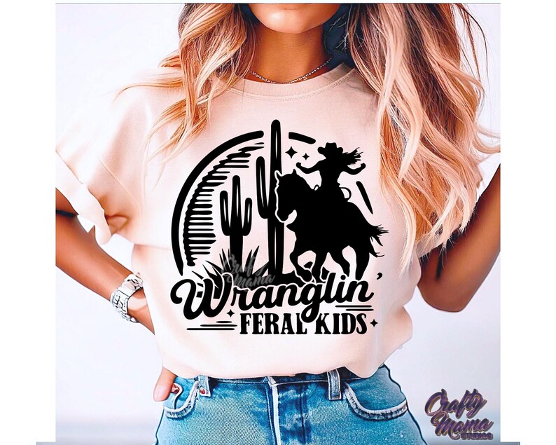 May include: A light peach-colored t-shirt featuring a black graphic of a cowgirl riding a horse, cacti, and the words "Wranglin' FERAL KIDS." The design is set within a semi-circle. The text "Crafty Mama Studios" is also visible.