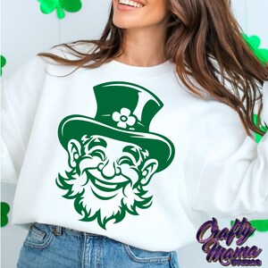 Pode incluir: Camisola branca com um gr&aacute;fico de um duende verde. O duende est&aacute; usando um chap&eacute;u verde com um trevo e tem um grande sorriso.