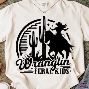 May include: Cream-colored t-shirt featuring a black graphic of a cowgirl riding a horse, with cacti and the words "Wranglin' Feral Kids." The design is in a Western style, suitable for casual wear.