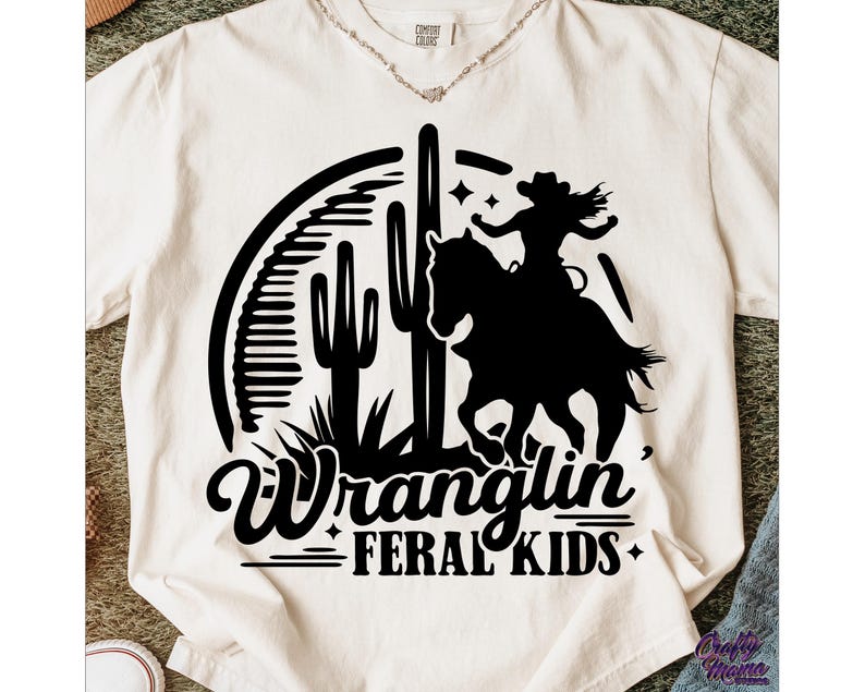 May include: Cream-colored t-shirt featuring a black graphic of a cowgirl riding a horse, with cacti and the words "Wranglin' Feral Kids." The design is in a Western style, suitable for casual wear.