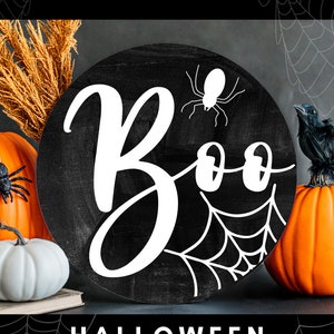 K&ouml;nnte beinhalten: Ein rundes Schild in Schwarzwei&szlig; mit dem Wort "Boo" in wei&szlig;er Kursivschrift. Im Hintergrund sind ein Spinnennetz und eine Spinne zu sehen. Das Schild ist von K&uuml;rbissen und Herbstdekoration umgeben.