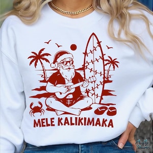 May include: White sweatshirt with a red graphic of Santa Claus playing a ukulele on a beach, with a surfboard, palm trees, and the words "MELE KALIKIMAKA". The design includes a crab, birds, and sandals.