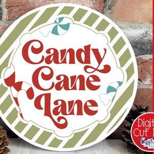 Op de afbeelding: Een rond, wit bord met een groene en witte gestreepte rand. Het bord zegt "Candy Cane Lane" in rode letters met een retro-lettertype. Het bord is versierd met snoepstokken en pepermuntjes.