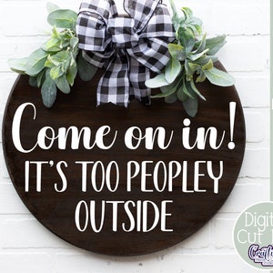 K&ouml;nnte beinhalten: Ein rundes Holzschild mit einer schwarz-wei&szlig;en karierten Schleife und Gr&uuml;n. Das Schild sagt "Come on in! It's too peopley outside."