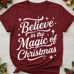 以下が含まれることがあります： 「Believe in the Magic of Christmas」の白い文字がプリントされた、バーガンディ色のTシャツ。デザインには星のアクセントが含まれています。このシャツは半袖で、クルーネックです。