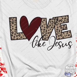 Puede incluir: Una camiseta blanca con un dise&ntilde;o de estampado de leopardo marr&oacute;n y negro que deletrea "LOVE" con un coraz&oacute;n rojo en el medio. El texto "like Jesus" est&aacute; escrito debajo de la palabra "LOVE".