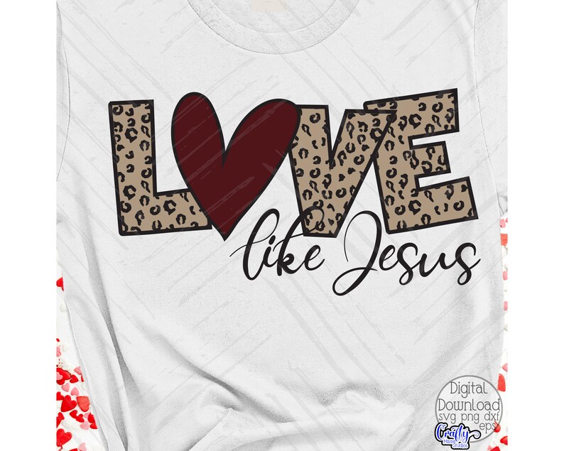 Puede incluir: Una camiseta blanca con un dise&ntilde;o de estampado de leopardo marr&oacute;n y negro que deletrea "LOVE" con un coraz&oacute;n rojo en el medio. El texto "like Jesus" est&aacute; escrito debajo de la palabra "LOVE".