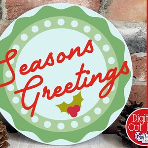 Op de afbeelding: Een rond, groen en wit kerstbord met een blauwe achtergrond. Het bord zegt "Seasons Greetings" in rode, cursieve letters met een groene hulsttak en rode bessen in het midden.