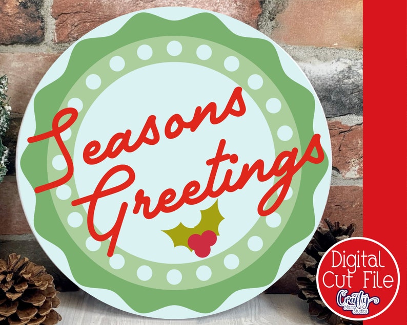 Op de afbeelding: Een rond, groen en wit kerstbord met een blauwe achtergrond. Het bord zegt "Seasons Greetings" in rode, cursieve letters met een groene hulsttak en rode bessen in het midden.
