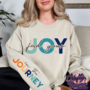 May include: A light beige sweatshirt with the text "find your JOY" in blue, green, and orange on the front. The left sleeve has the text "in the JOURNEY" in orange, blue, and green.