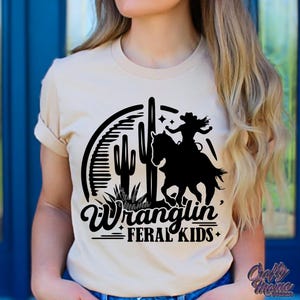 May include: Beige t-shirt featuring a black graphic of a person riding a horse, cacti, and the words "Wranglin' Feral Kids." The shirt has short sleeves and a crew neck.